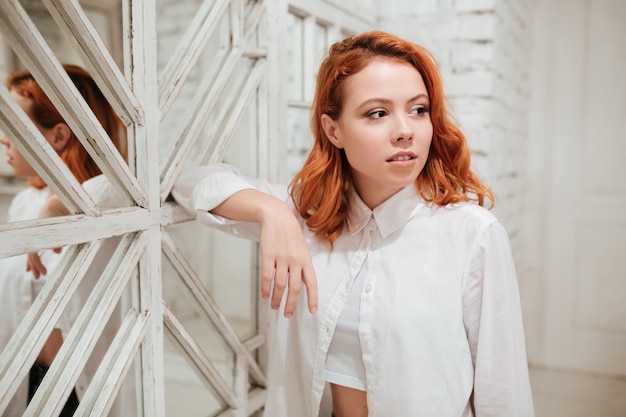 Retrato de mujer joven hermosa pelirroja vestida con camisa blanca de pie junto al espejo en la cafetería.