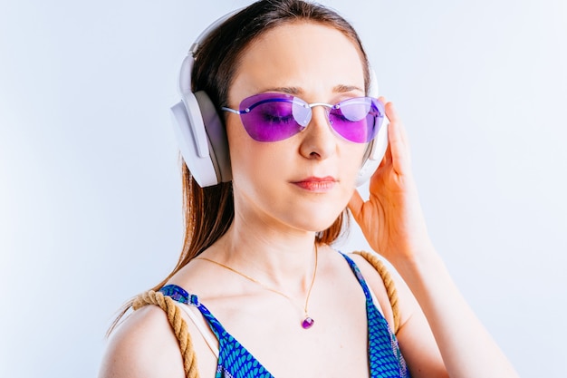 Retrato de mujer joven hermosa sobre fondo blanco con gafas de verano escuchando música con auriculares inalámbricos. concepto ir de vacaciones