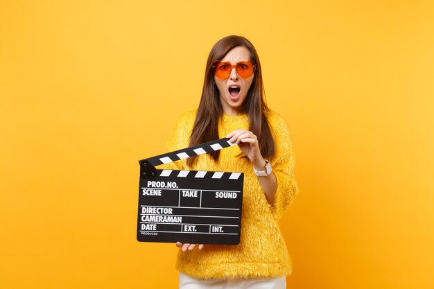 Retrato de mujer joven irritada en gafas de corazón naranja suéter de piel sosteniendo claqueta de cine negro clásico aislado sobre fondo amarillo. Estilo de vida de las emociones sinceras de la gente. Área de publicidad.