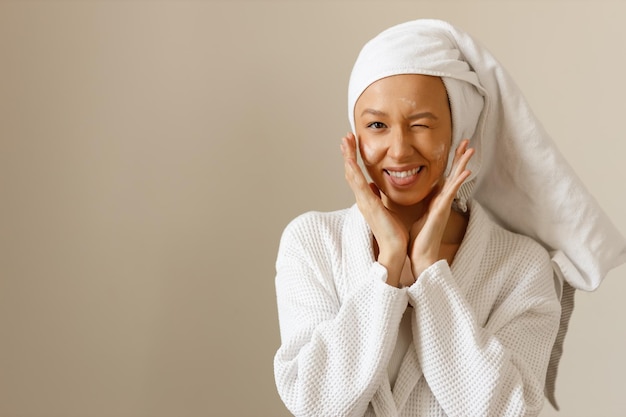 Retrato de mujer joven juguetona en bata blanca y toalla en la cabeza aplicando loción o crema hidratante en la cara Cuidado facial profesional después de lavar Ojos guiñando Fondo aislado