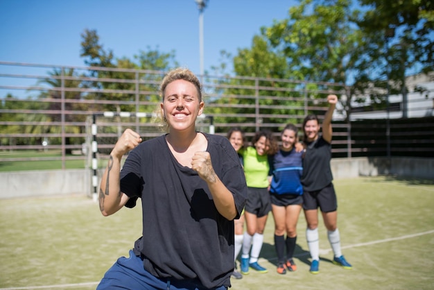 Retrato de mujer joven juguetona en el campo de fútbol. Deportista con uniforme oscuro guiñando un ojo a la cámara, alegres compañeros de equipo en segundo plano. Deporte, ocio, concepto de estilo de vida activo.