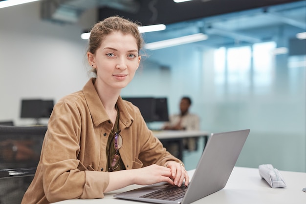 Retrato de mujer joven con laptop sonriendo a la cámara en la oficina o biblioteca escolar, espacio de copia
