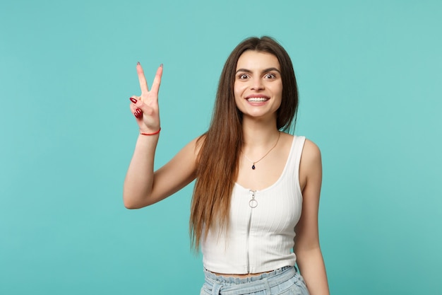 Retrato de mujer joven linda sonriente en ropa casual ligera que muestra el signo de la victoria aislado sobre fondo de pared azul turquesa en estudio. Personas sinceras emociones, concepto de estilo de vida. Simulacros de espacio de copia.