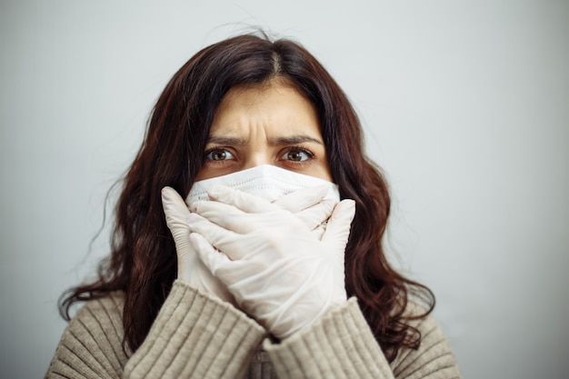 El retrato de una mujer joven se lleva las manos a la boca mostrando el signo de silencio y se queda en casa durante la cuarentena debido a la pandemia de coronavirus. Hermosa chica con guantes y mascarilla médica. Epidemia de Covid-19.