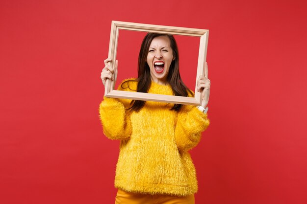 Retrato de mujer joven loca en suéter de piel amarilla gritando, sosteniendo el marco de imagen aislado sobre fondo rojo brillante de la pared en el estudio. Personas sinceras emociones, concepto de estilo de vida. Simulacros de espacio de copia.