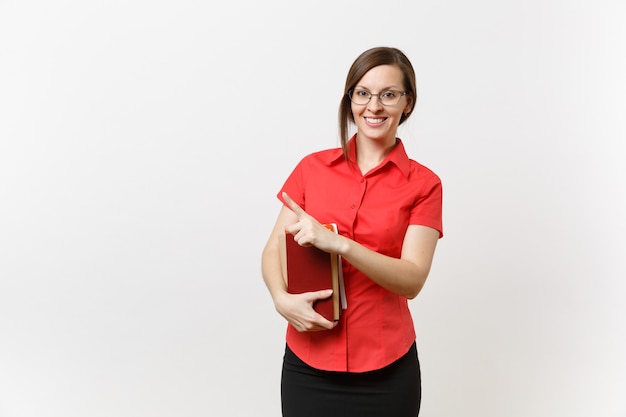 Retrato de mujer joven maestra en camisa roja, falda y gafas sosteniendo libros, señalando con el dedo a un lado en el espacio de la copia aislado sobre fondo blanco. Educación o enseñanza en el concepto de universidad de secundaria.