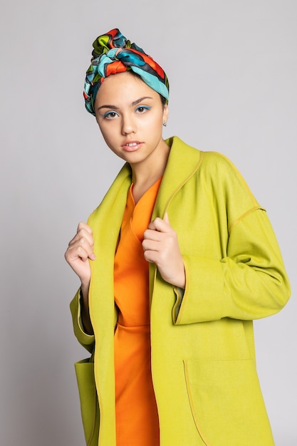 Retrato de una mujer joven con maquillaje brillante y un pañuelo de moda. Fondo claro. Belleza, moda, concepto de maquillaje. chica con un abrigo verde brillante, vestido naranja brillante.