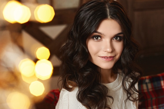 Retrato de una mujer joven con maquillaje de moda posando con luces navideñas en el fondo.