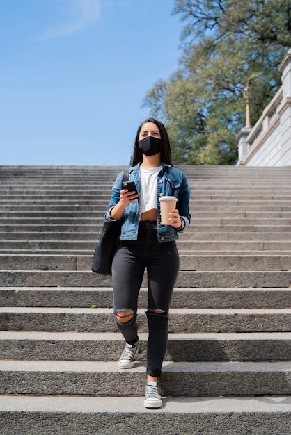 Retrato de mujer joven con máscara facial y usando su teléfono móvil mientras camina al aire libre. Nuevo concepto de estilo de vida normal. Concepto urbano.