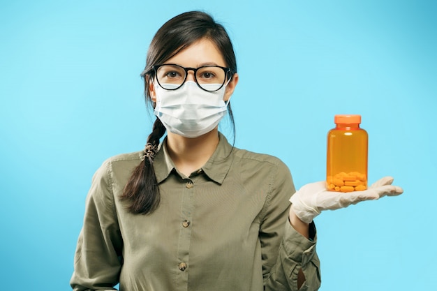 Retrato de una mujer joven en máscara médica protectora y guantes sosteniendo una botella naranja con pastillas o vitaminas en azul.