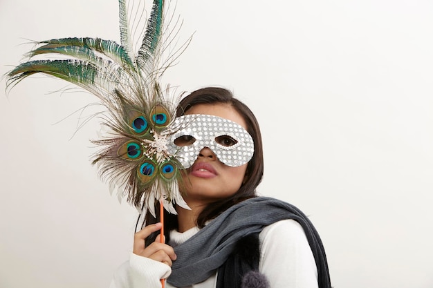 Retrato de una mujer joven con una máscara para los ojos sosteniendo plumas de pavo real contra un fondo blanco