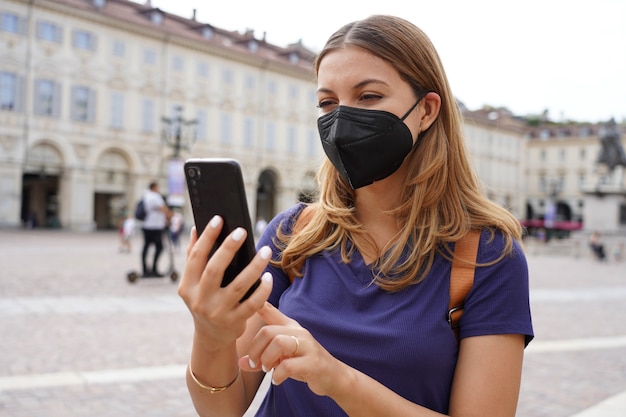 Retrato de mujer joven con máscara protectora negra FFP2 KN95 mediante teléfono móvil con fondo de ciudad