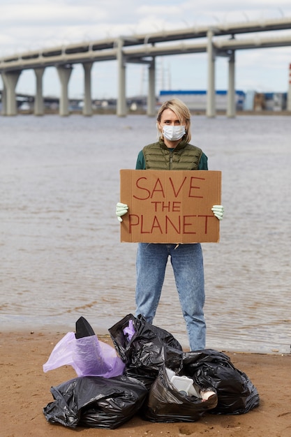 Foto retrato de mujer joven con máscara protectora sosteniendo cartel salvar el planeta mientras está de pie cerca de la costa con bolsas de basura