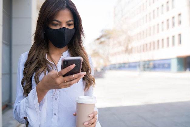 Retrato de mujer joven con máscara protectora y usando su teléfono móvil mientras está de pie al aire libre en la calle. Nuevo concepto de estilo de vida normal. Concepto urbano.