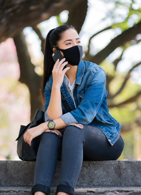 Retrato de mujer joven con mascarilla y hablando por teléfono mientras está sentado en las escaleras al aire libre