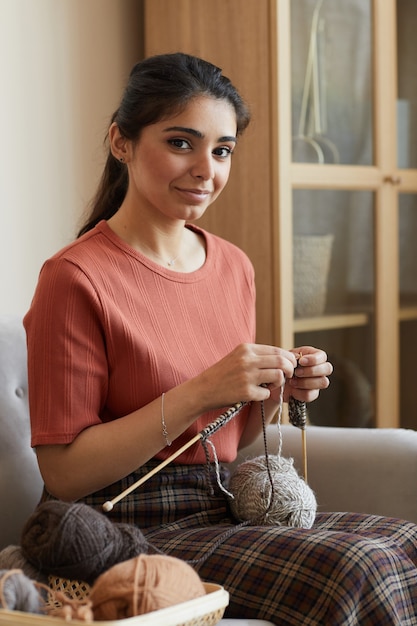 Retrato de mujer joven mirando al frente mientras teje la ropa de lana sentado en el sofá en casa