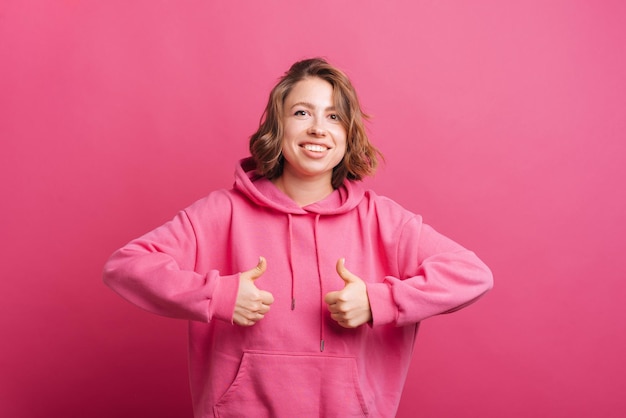 Retrato de mujer joven de moda en sudadera con capucha rosa mostrando los pulgares hacia arriba y sonriendo a la cámara