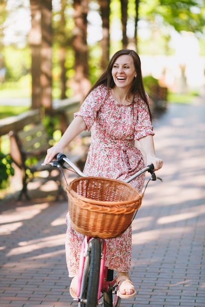 Retrato de mujer joven de moda en vestido largo de flores rosa cabalgando por el callejón en bicicleta vintage con canasta para compras, comida o flores al aire libre, tiempo de recreación femenina hermosa en el parque de primavera o verano.