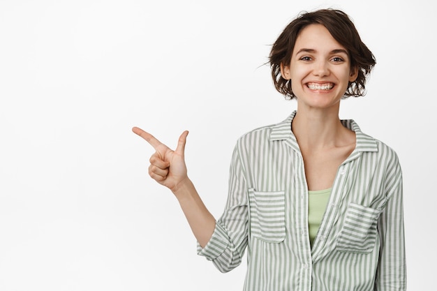 Retrato de mujer joven moderna, emprendedora apuntando con el dedo a la izquierda, mostrando publicidad y sonriendo, de pie sobre blanco