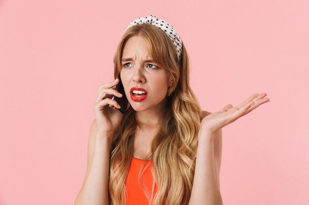 Retrato de mujer joven molesta con pelo largo y rizado frunciendo el ceño y hablando por teléfono inteligente aislado sobre pared rosa