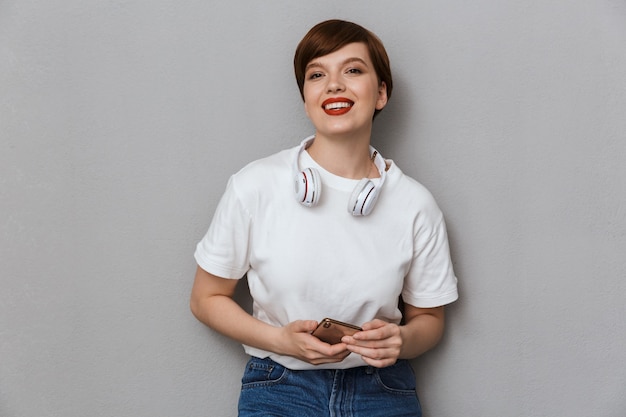 Retrato de mujer joven morena con auriculares sosteniendo celular aislado sobre pared gris