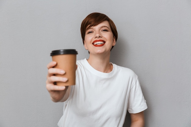 Retrato de mujer joven morena con camiseta casual sonriendo y sosteniendo la taza de café para llevar aislado sobre pared gris