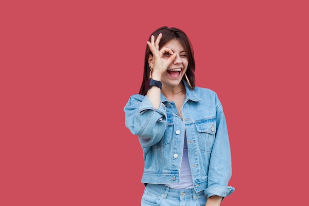 Retrato de mujer joven morena hermosa feliz divertida con maquillaje en estilo casual de mezclilla de pie con el signo de Ok en sus ojos y mirando a cámara. tiro del estudio de interior, aislado en fondo rojo.