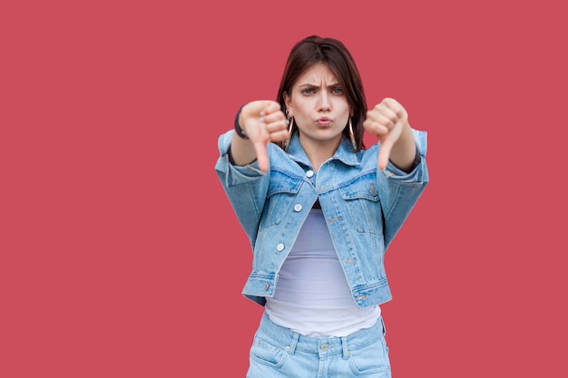 Retrato de mujer joven morena hermosa insatisfecha con maquillaje en denim estilo casual de pie, pulgares hacia abajo y mirando a cámara con disgusto. tiro del estudio de interior, aislado en fondo rojo.