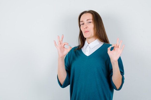 Retrato de mujer joven mostrando gesto ok en suéter sobre camisa blanca y mirando complacido vista frontal