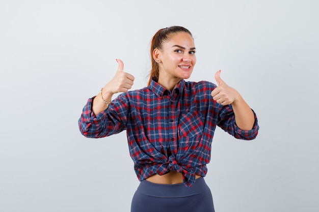 Retrato de mujer joven mostrando los pulgares para arriba en camisa a cuadros, pantalones y mirando feliz vista frontal