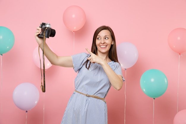 Retrato de mujer joven muy feliz en vestido azul haciendo selfie dedo índice señalando en cámara de fotos vintage retro sobre fondo rosa pastel con globo de aire colorido. Concepto de fiesta de cumpleaños.