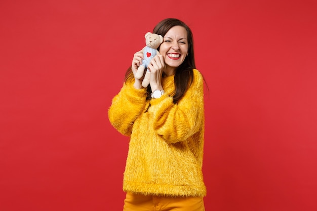 Retrato de mujer joven muy sonriente en suéter de piel amarilla mantenga peluche oso de peluche aislado sobre fondo de pared rojo brillante en estudio. Personas sinceras emociones, concepto de estilo de vida. Simulacros de espacio de copia.