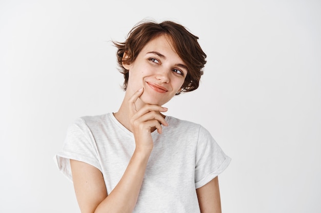 Retrato de mujer joven natural con peinado corto, mirando pensativo a un lado, sonriendo y soñando con algo, de pie pensativo en la pared blanca