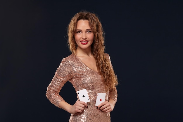 Retrato de una mujer joven o de cabello castaño sonriendo, sosteniendo un par de ases con un vestido de cóctel dorado sobre fondo negro. Concepto de casino, industria del juego