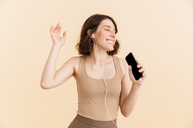 Retrato de mujer joven optimista vestida con ropa casual cantando mientras escucha música con auriculares y teléfono celular aislado