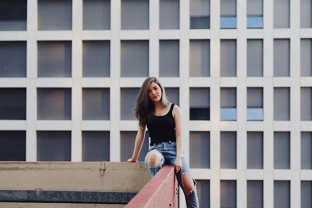 Retrato de una mujer joven con pantalones vaqueros rasgados sentada en la barandilla contra un edificio moderno