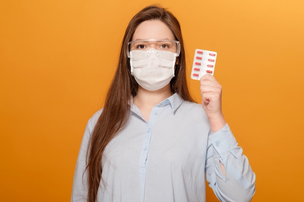 Retrato de mujer joven en la pared de color naranja en la máscara médica protectora con pastillas en la mano