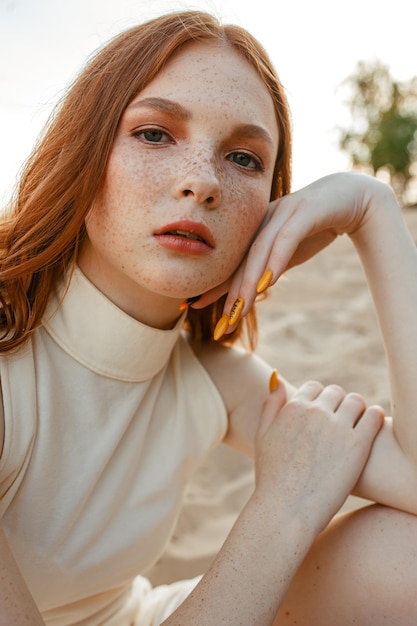 Retrato de mujer joven pelirroja con pecas tocando la mejilla y mirando a la cámara en la naturaleza