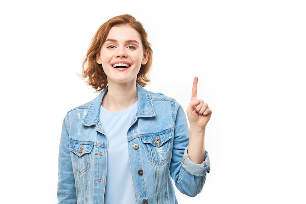 Retrato de mujer joven pelirroja positiva en jeans casuales sonriendo señalando con el dedo el espacio de copia vacío para texto o producto aislado en banner publicitario de fondo blanco