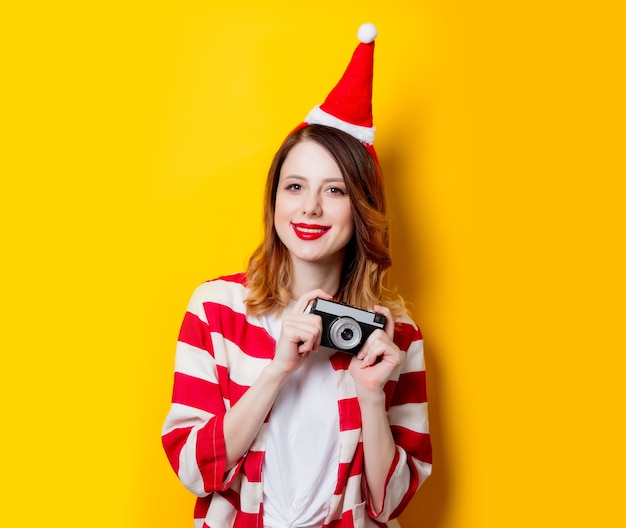 Retrato de mujer joven pelirroja con sombrero de Santa Claus y camisa a rayas con cámara clásica sobre fondo amarillo. tiempo de Navidad