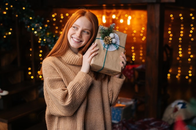 Retrato de mujer joven pelirroja sonriente agitando la caja de regalo con regalos de Navidad envueltos