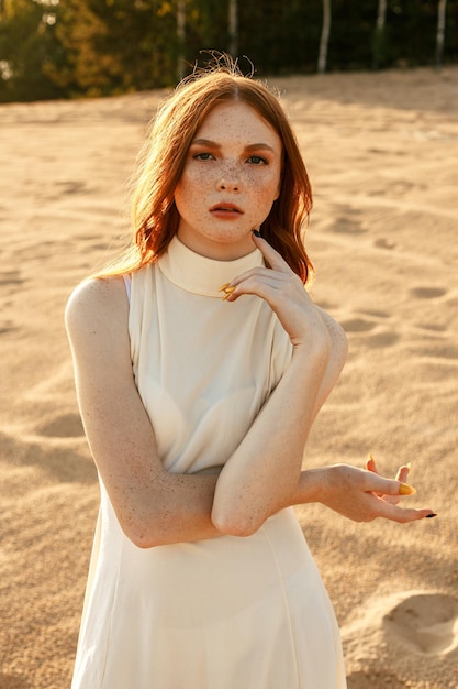 Retrato de mujer joven de pelo jengibre con vestido de verano mirando a la cámara en la playa de arena