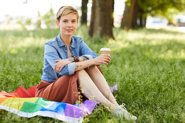 Retrato de mujer joven con pelo rubio corto sonriendo mientras está sentado en el césped y tomando café en el parque