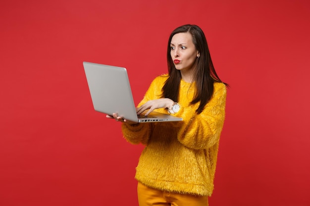 Retrato de mujer joven perpleja confundida en suéter de piel amarilla con ordenador portátil aislado sobre fondo rojo brillante de la pared en el estudio. Concepto de estilo de vida de emociones sinceras de personas. Simulacros de espacio de copia.