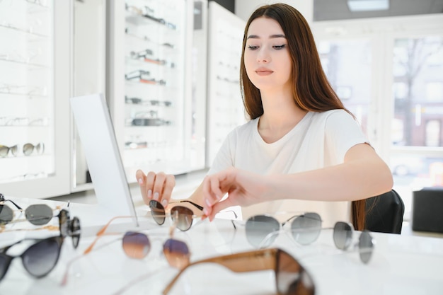 Retrato de una mujer joven de pie en la tienda y probando gafas de sol cerca de un espejo