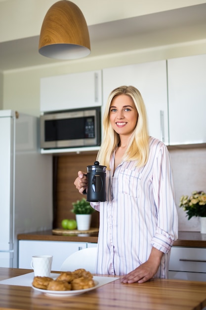 Retrato de mujer joven en pijama con prensa de café francés de pie en la cocina moderna