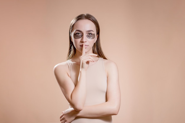 Retrato de mujer joven posando con parches de belleza negros aplicados debajo de los ojos sobre beige.