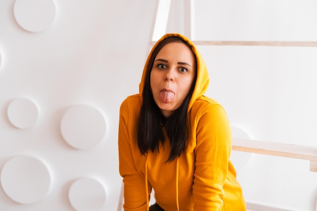 Retrato de mujer joven posando sobre fondo blanco. Bonita morena con capucha amarilla cerca de la escalera de tijera en el fondo de la pared blanca.