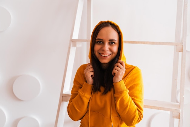 Retrato de mujer joven posando sobre fondo blanco. Bonita morena con capucha amarilla cerca de la escalera de tijera en el fondo de la pared blanca.