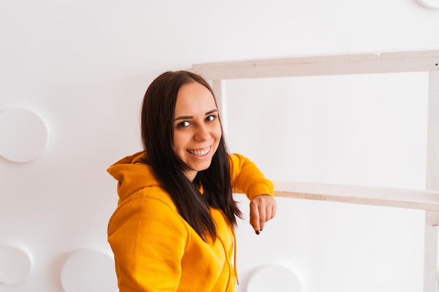 Retrato de mujer joven posando sobre fondo blanco. Bonita morena con capucha amarilla cerca de la escalera de tijera en el fondo de la pared blanca.